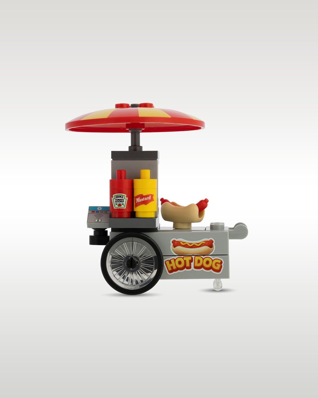 Side close-up view of the Trader Bricks custom toy brick hot dog stand, highlighting the large bicycle-style wheels, built-in grill area, and printed hot dog sign