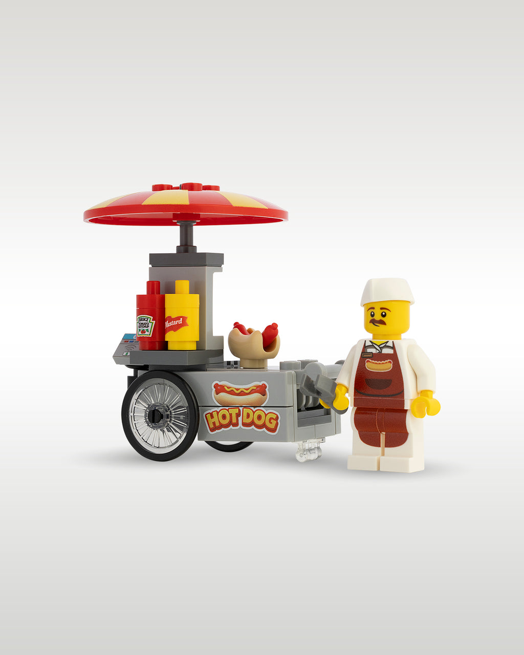 Custom brick-built Hot Dog stand food cart MOC set by Trader Bricks, featuring a red and yellow umbrella, custom printed Hot Dog sign, condiment bottles, and a street food vendor minifigure