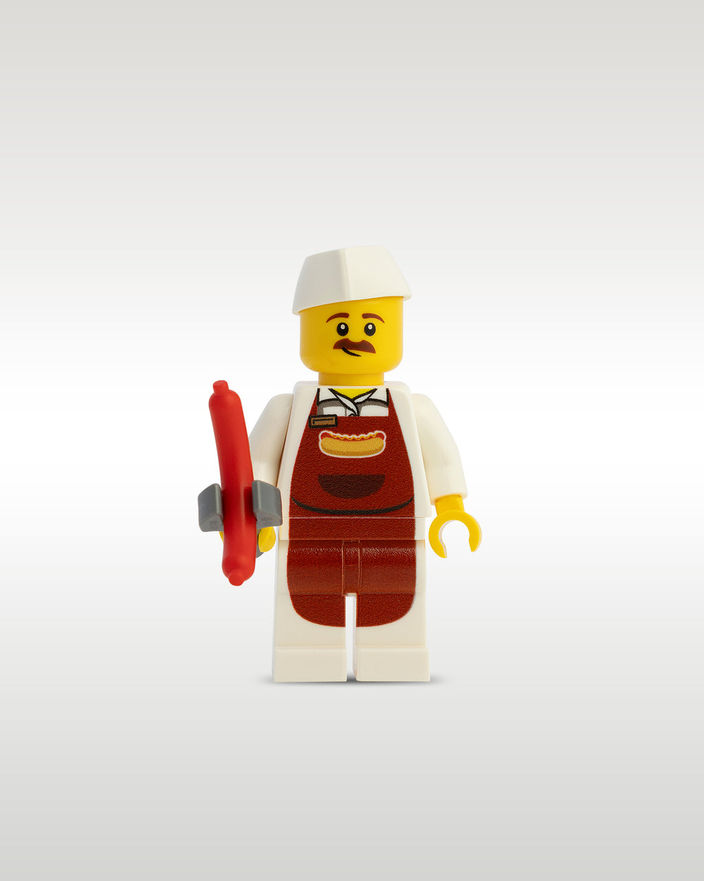 Custom printed street food vendor minifigure by Trader Bricks, featuring a mustache, a white chef uniform with a brown apron, a printed hot dog graphic on the torso, and a white service cap